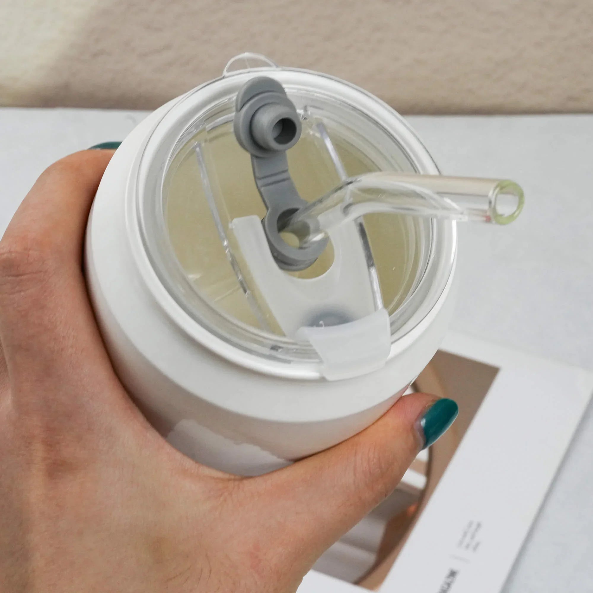 Hand holding a white tumbler with a clear lid and straw on a light background